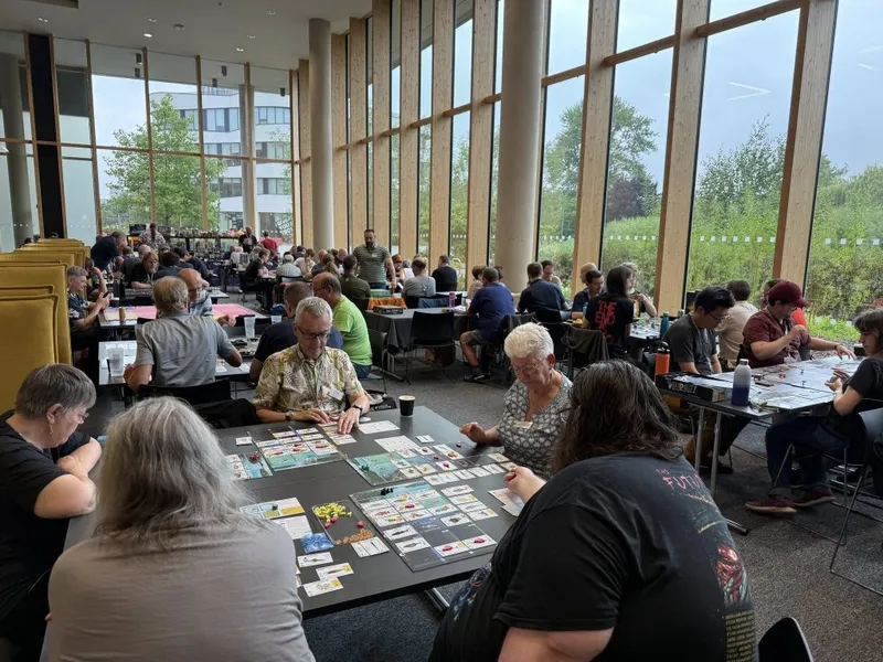 A bustling board game convention scene at the University of Northampton, featuring numerous tables filled with players engaged in various games. In the foreground, a group of four players is focused on a game spread across their table, with colorful game components and cards visible. Large windows in the background allow natural light to fill the space, highlighting the many attendees seated at tables throughout the room.