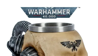 Beige ceramic mug shaped like a stylized human skull with mechanical augmentations. A metal rim lines the top of the mug. Two large, cylindrical, metallic mechanical components resembling pistons or servos are attached to the sides of the skull, connected by coiled black cables. A black eagle emblem with outstretched wings and a skull in the center is tattooed or printed on the forehead. The background shows a metallic banner with the text "WARHAMMER 40,000" in bold, futuristic lettering. The banner has a red and white stripe along the bottom edge.