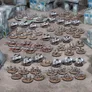 A large array of 10mm-scale miniatures is displayed on a tabletop, showcasing a sci-fi wargaming scene. The scene features various types of vehicles and infantry, including sleek hovercraft and armored soldiers, all set against a backdrop of modular, futuristic buildings. The miniatures are arranged in organized formations, highlighting the two distinct armies, with a mix of colors primarily in shades of gray and metallic tones.