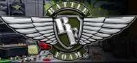 Stylized emblem featuring a shield design. At the top of the shield, bold white capital letters spell "BATTLE". In the center, a large white number "3" is integrated with a sword pointing downward. Below the shield, a green banner with white capital letters reads "FOAM" and to the right of it, a registered trademark symbol "(R)". The background depicts a cluttered tabletop with miniature terrain pieces, small figures, dice, and a red vehicle model. The setting appears to be a gaming or hobby workspace.