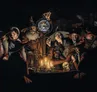 Group of robed figures gathered around a glowing globe, candles, and a skull on a table.