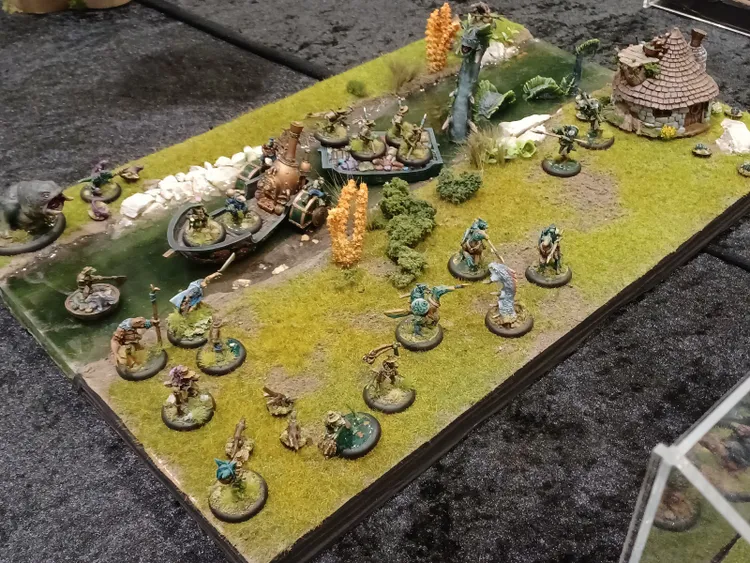 A detailed tabletop diorama showcases a vibrant fantasy scene featuring various miniatures on a grassy terrain. In the foreground, several anthropomorphic figures, including reptilian warriors and other fantastical creatures, are positioned strategically, while a small boat is docked along a waterway. The backdrop includes lush vegetation, a serpentine creature, and a rustic structure, all contributing to the immersive world of Anyaral.