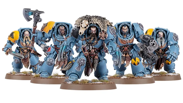 A trio of Space Wolves miniatures stands prominently, showcasing their detailed armor and weaponry. The central figure, likely Logan Grimnar, is adorned in blue terminator armor with a fur cloak and a skull, wielding an axe. Flanking him are two lieutenants: one with a large weapon and intricate details, and the other featuring a distinctive beard and additional armor embellishments, all set against a base with tufts of grass and snow.