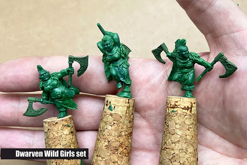 Three small, green miniatures from the "Dwarven Wild Girls" set are being held in a hand, each perched on corks. The miniatures depict stout female dwarves, each wielding an axe and showcasing distinct poses and expressions. The background is a neutral beige, allowing the details of the miniatures to stand out prominently.