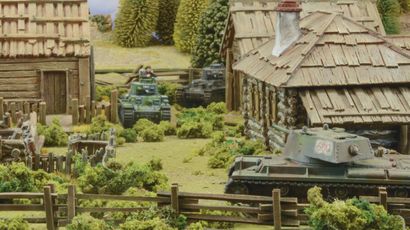 A detailed tabletop scene features several miniature tanks amidst a rural setting, including a wooden cabin and lush greenery. In the foreground, a tank with the number "600" is positioned near a wooden fence, while another tank is seen further back with a crew member visible. The background includes trees and additional structures, creating a vibrant battlefield atmosphere.