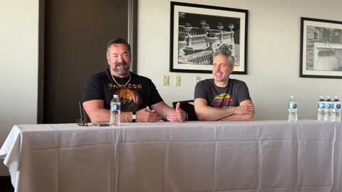 Two men are seated at a table during a panel discussion. On the left, a man with a beard, wearing a black shirt featuring a colorful graphic and a necklace, gestures with his hands while speaking. On the right, a man with short gray hair, dressed in a gray t-shirt with a retro design, smiles and listens attentively. Behind them, a framed black-and-white photograph of a decorative railing is visible on the wall. A bottle of water and a small device are placed on the table in front of them.
