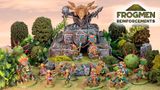 A vibrant scene showcases a variety of colorful Space Frogmen miniatures, including a frogman chieftain and cavalry, positioned around a rocky outcrop adorned with ancient symbols and a large decorative skull. Lush greenery and spiky plants surround the figures, enhancing the fantastical atmosphere. The text "FROGMEN REINFORCEMENTS" is prominently displayed in the corner, indicating the theme of the Kickstarter campaign.