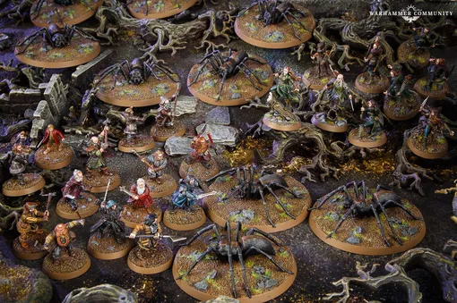 A detailed tabletop scene features various miniatures from the Middle-earth Strategy Battle Game, depicting a tense moment in Mirkwood. Prominent are several large spiders on circular bases, surrounded by a mix of characters including Bilbo, Thorin, and other dwarves, all positioned on a textured terrain with rocks and twisted roots. The dark, earthy colors of the setting enhance the atmosphere of the iconic "Flies and Spiders" chapter from The Hobbit.