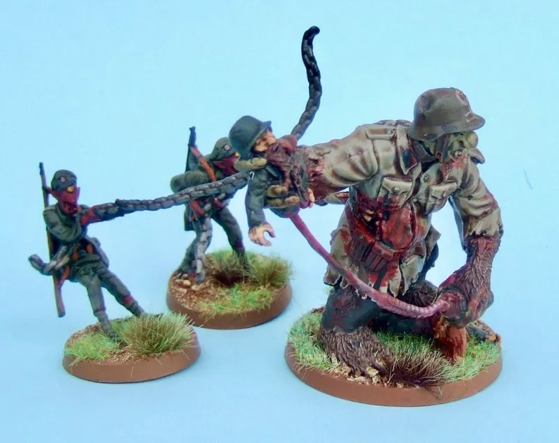 A trio of miniatures from the Panzerfäuste game is depicted, featuring two soldiers in dark uniforms and masks, each holding a weapon, and a larger, grotesque figure resembling a zombie or monstrous soldier. The larger figure has a tattered uniform and is being restrained by a long, tentacle-like appendage. The bases of the miniatures are painted in earthy tones with tufts of grass, set against a light blue background.