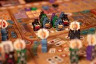 Eight black cat-themed devil miniatures playing drums, standing on a wooden board with a path and buildings. Two fire-breathing devil figures with flames. Scattered cards with symbols and numbers.