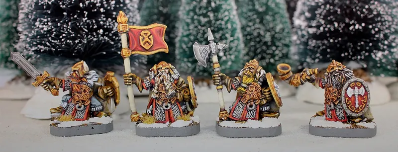 Three painted miniature figures of fantasy dwarves stand on round bases in a snowy outdoor setting with blurred evergreen trees in the background.

The left figure wears a horned helmet, patterned tunic, and fur cloak, holding a pole-mounted red banner with a gold emblem of a snarling face. The figure also carries a round shield on their back.

The center figure, slightly behind the others, wears a helmet with a nose guard, a fur-lined cloak, and carries a pole weapon with a curved blade.

The right figure, holding a large double-bladed axe over one shoulder, wears a horned helmet, fur cloak, and patterned tunic. Their beard is braided and wrapped around their waist.