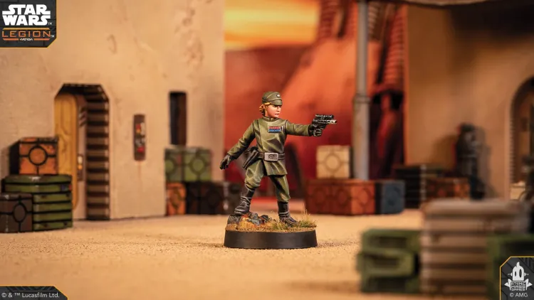 A detailed miniature of a customizable Imperial Officer stands prominently in the foreground, wearing a green uniform with a rank insignia and a cap. The background features a sandy terrain with crates and a building entrance, suggesting a desert environment typical of the Star Wars universe. The scene is set within a tabletop gaming context, enhancing the immersive experience for players.