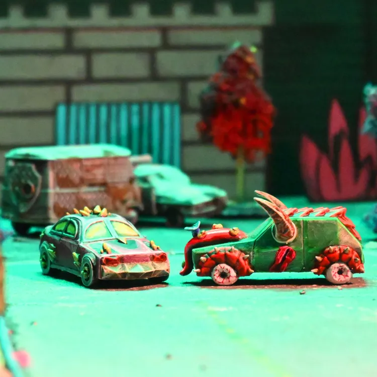 Two uniquely designed miniature vehicles are positioned prominently in the foreground, showcasing their vibrant colors and intricate details. The left vehicle is a sleek, dark car adorned with spikes and a stylized front, while the right vehicle features a more rugged design with a green body, red accents, and menacing horns. The background includes blurred outlines of additional vehicles and a stylized urban environment, enhancing the chaotic atmosphere of the racing game.