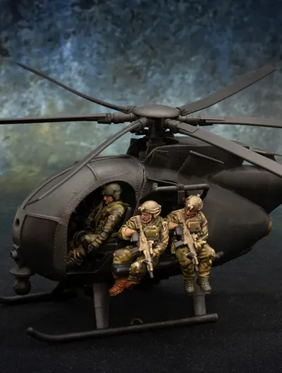 A detailed model of a Little Bird helicopter, featuring a dark color scheme, is prominently displayed. Two miniature soldiers are seated on the helicopter's skids, equipped with rifles, while another soldier is visible inside the cockpit. The background features a textured gradient, enhancing the focus on the helicopter and its crew.