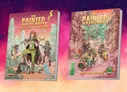 Two books titled The Painted Wastelands: left cover shows a central female character with a staff, surrounded by desert ruins, a cat, and two other figures; text reads "Adventurer’s Handbook." Right cover depicts a fantasy creature atop ruins with a green-tinted desert; text reads "Campaign Guide."
