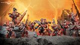 A group of World Eaters miniatures stands prominently against a fiery orange background, depicting a chaotic battlefield. The figures are heavily armored, predominantly in red with black and silver details, showcasing various weapons and intricate designs, including a large, imposing character at the center. The rocky terrain beneath them adds to the dramatic atmosphere, emphasizing their fierce presence in the Warhammer 40k universe.