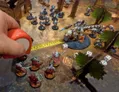 A tabletop gaming scene features a vibrant battlefield with various miniatures positioned across a textured gaming mat. In the foreground, a player is using a measuring tape to gauge distance between a group of robotic figures in red capes and a large, dinosaur-like creature with a rider, which stands on a circular base. Surrounding them are additional miniatures, terrain pieces, and several white dice scattered on the ground, creating an immersive gaming environment.