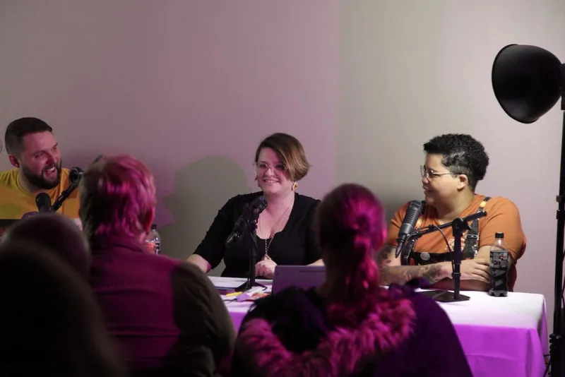 Four people at a table with microphones, one speaking, pink and purple lighting.
