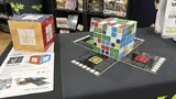 A wooden cube puzzle game on a black table with six colored cube types—forest, ocean, cave, desert, lava, tundra—partially assembled. Two game boards with numbered tiles and a red scoring token lie nearby. Shelves in background display other board game boxes.