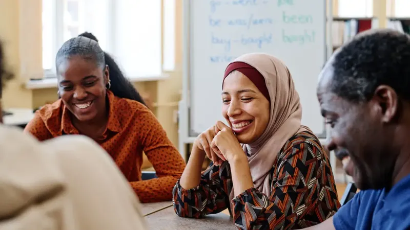 A group of individuals is engaged in a conversation around a table, with a focus on a woman wearing a light-colored hijab and a patterned blouse, who is smiling warmly. To her left, a man with a dark complexion and gray hair is also smiling, while another person, partially visible, wears a rust-colored shirt. In the background, a whiteboard displays written words, suggesting an educational or collaborative setting.