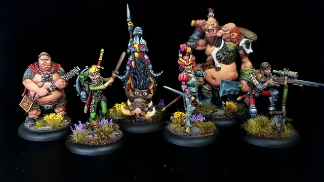 A group of eight intricately painted Moonstone miniatures stands against a black background. The characters include a muscular figure with a weapon, a green-skinned goblin in colorful attire, a knight in armor, and various other fantastical beings, each displaying unique features and vibrant colors. They are positioned on circular bases adorned with tufts of grass and small flowers, showcasing the detailed artistry of the miniatures.