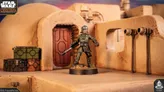A detailed miniature of a character stands in front of a rounded, tan structure resembling a building from the Star Wars universe, with a red door and various mechanical elements visible. The character is dressed in a green uniform, holding a weapon, and is positioned on a rocky base. Nearby, there are several colorful crates in green, orange, and black, adding to the scene's immersive atmosphere.