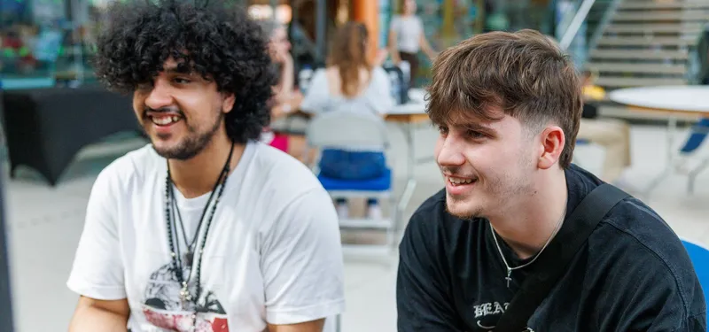 Two young men are seated in a casual setting, engaged in conversation. One has curly hair and is wearing a white t-shirt, while the other has shorter hair and is dressed in a black t-shirt. In the background, people are gathered around tables, with some seated and others standing, creating a lively atmosphere typical of a gaming event.