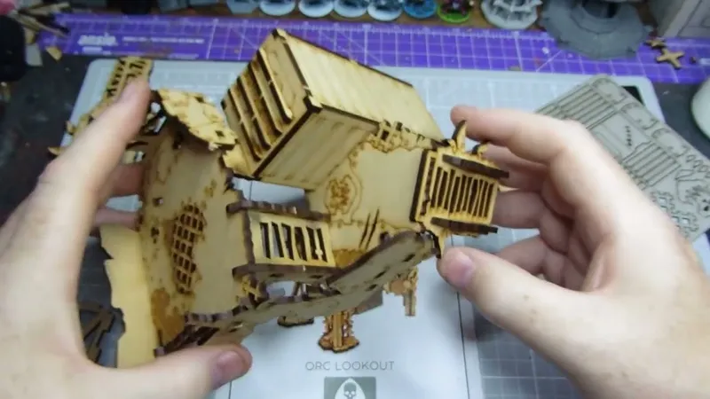 A partially assembled Orc Lookout tower made of laser-cut wood is being held by two hands. The structure features multiple levels, with a slanted roof and wooden slats, showcasing intricate details like windows and a skull motif. In the background, a crafting mat and various miniatures are visible, indicating a tabletop gaming environment.