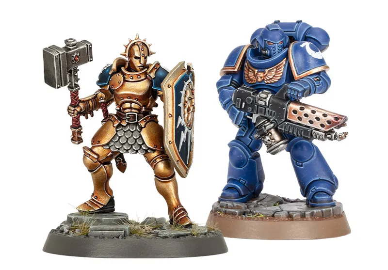 A golden-armored Stormcast Eternals Liberator stands on a rocky base, wielding a large hammer in one hand and a shield emblazoned with a sun motif in the other. Next to it, a blue-armored Infernus Space Marine holds a firearm, showcasing intricate details on both miniatures. The base features tufts of grass and rocky terrain, enhancing the overall display of these Warhammer figures.