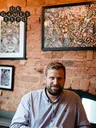 Man in light blue shirt holding a glass, brick wall with framed colorful artwork behind.