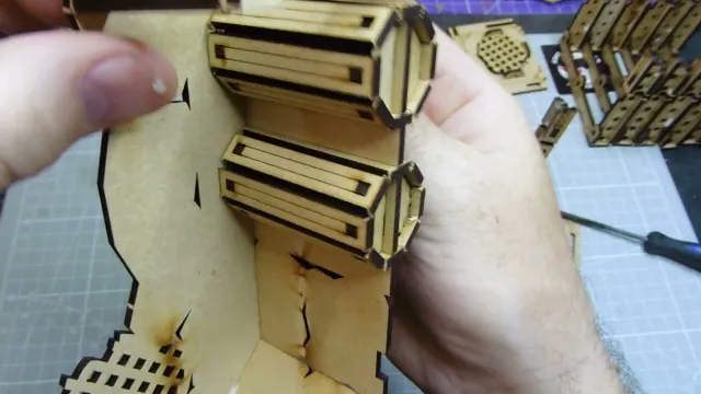 A close-up view of a partially assembled Orc Mega Bunker model from TT Combat, specifically the Orc Sektor section. The image shows a hand holding a piece of the model, which features intricate cut-out designs and layered components. The background includes a grid pattern, suggesting a crafting or modeling workspace.