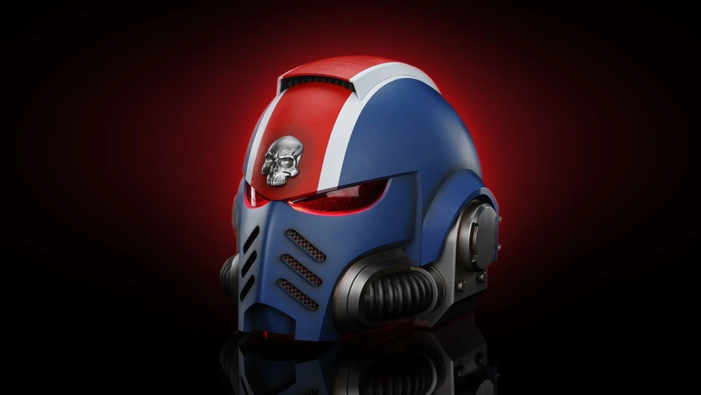 A wearable Space Marine helmet features a striking design with a blue and red color scheme, accented by a prominent skull emblem on the forehead. The helmet has a sleek, angular shape with dark eye lenses and ventilation grills on the front. It is set against a dramatic dark red background, enhancing its imposing appearance.