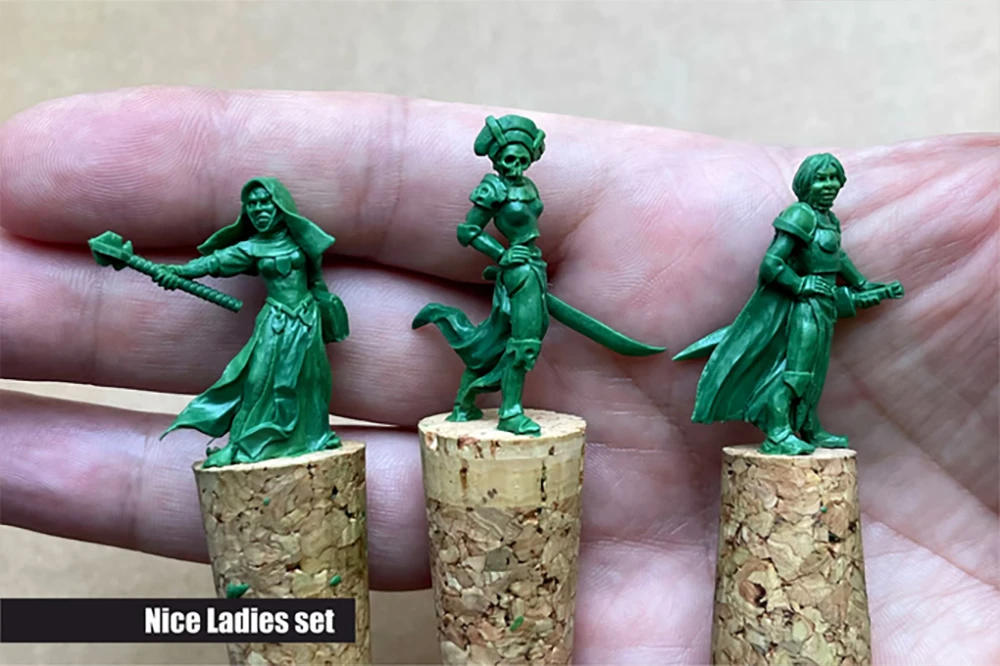 Three green resin miniatures are displayed on cork bases, held in a hand. The figures represent fantasy female characters: on the left, a warrior wielding a mace and wearing a flowing robe; in the center, a character with a sword and a distinctive headdress; and on the right, another figure in a cloak. The text 