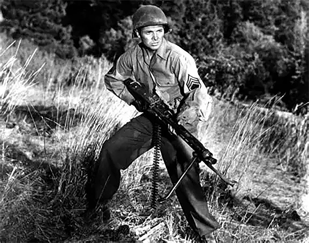 A black-and-white image features a soldier in a military uniform, wearing a helmet and a sergeant's insignia, crouched in a grassy field. He holds a machine gun with both hands, poised as if ready for action. The background consists of blurred greenery, suggesting a natural setting.