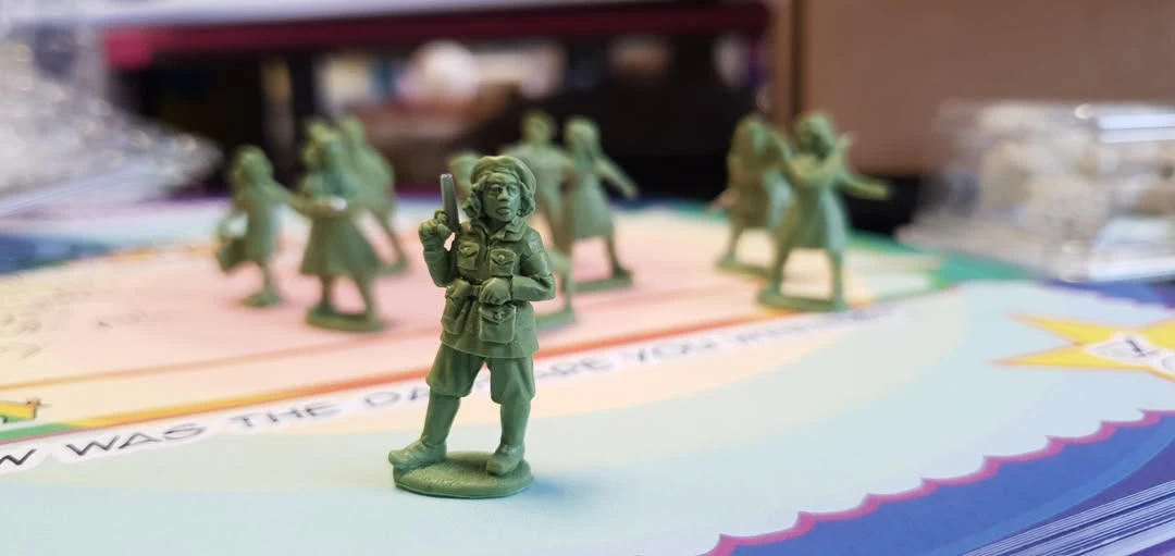 A detailed miniature figure of a woman stands prominently in the foreground, holding a pistol and wearing a military-style uniform, complete with a belt and pockets. Behind her, several other smaller green miniatures depict women in various poses, suggesting a scene of resistance. The background features a colorful tabletop surface with text, adding context to the setting.