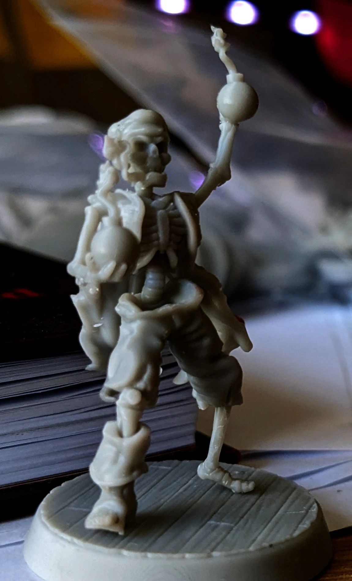 A detailed miniature of a skeleton pirate stands with one arm raised, holding a round object, possibly a cannonball or a drink. The figure is intricately sculpted, showcasing skeletal features and pirate attire, including tattered clothing. The background is slightly blurred, revealing stacks of game-related materials, suggesting a tabletop gaming environment.