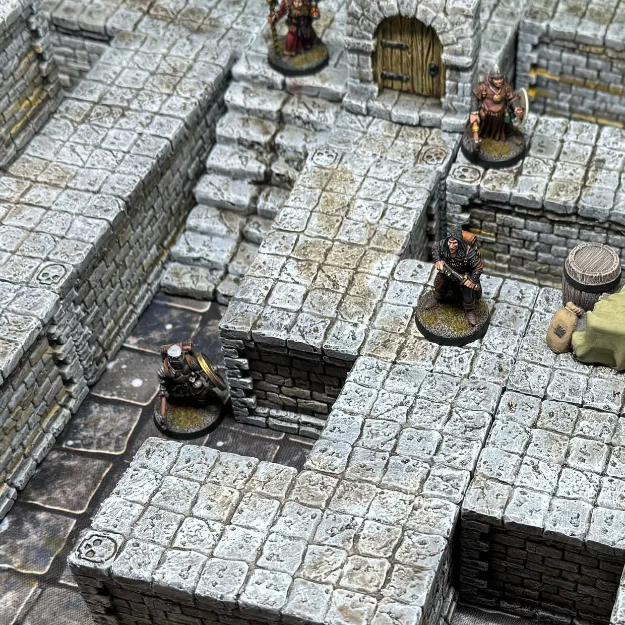 A detailed tabletop scene features modular dungeon blocks made of textured stone, arranged to create walls, platforms, and staircases. Several miniatures, including two armored figures and a robed character, are positioned on the blocks, exploring the dungeon layout. A wooden barrel and a small pile of supplies are visible, adding to the immersive environment.
