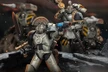 A squad of Iron Warriors action figures is prominently displayed, showcasing their detailed designs and articulation. The foreground features a Sergeant wielding a power sword and a plasma pistol, while a command squad member stands behind, holding a staff. The figures are set against a dramatic backdrop, enhancing their imposing presence with hints of smoke and debris.
