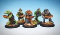 A group of four intricately painted orc miniatures stands against a light blue background. Each orc is armed with various hand weapons, showcasing a mix of colors and details in their armor and features. The miniatures are positioned on round bases adorned with small tufts of grass and skulls, highlighting their dynamic poses and unique characteristics.
