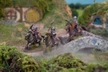 Three mounted halfling cavalry figures are depicted, each riding stout ponies and equipped with spears. The riders wear armor and helmets, showcasing a mix of colors, including silver and red accents. The scene is set in a lush, grassy landscape with rocky terrain and a distant background of rolling hills, creating a vibrant and dynamic atmosphere.