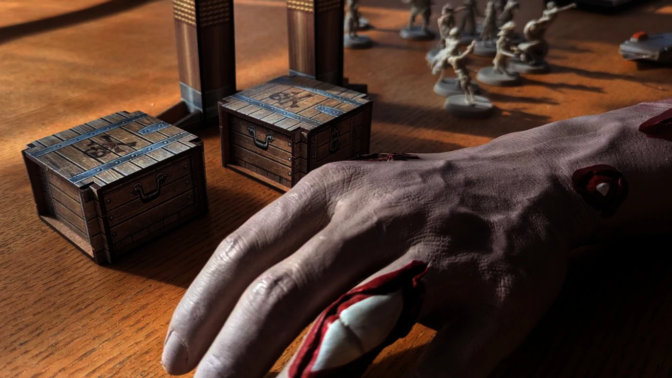 A close-up view of a tabletop scene featuring two small, wooden treasure chests with metal accents, each adorned with pirate-themed symbols. In the background, a tall, wooden structure resembling a ship's mast is partially visible, along with a human hand resting on the table, suggesting engagement with the game. The warm wooden surface adds to the overall pirate aesthetic of the setup.