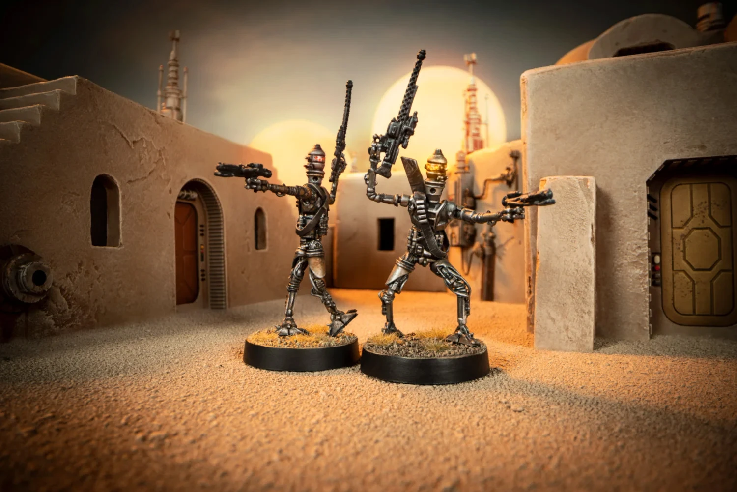 Two IG-Series Assassin Droids stand in a sandy, desert-like environment, each on a round black base. The droids are metallic with intricate detailing, one holding a blaster in each hand, while the other raises a weapon high. The background features a stylized, sci-fi setting with structures and soft lighting, evoking a Star Wars atmosphere.