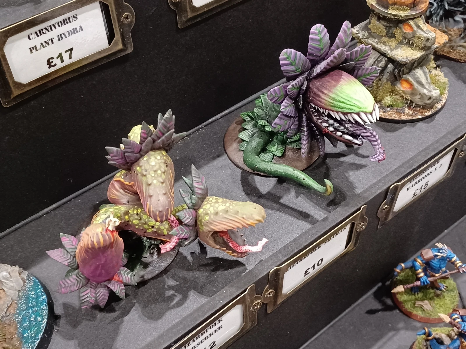 A display of intricately painted fantasy miniatures features several man-eating plants, showcasing vibrant colors and detailed designs. The plants have multiple heads, with some exhibiting bright greens and purples, while others display a mix of pinks and yellows. The miniatures are arranged on a dark surface, with price tags visible next to them, highlighting their unique and imaginative designs.