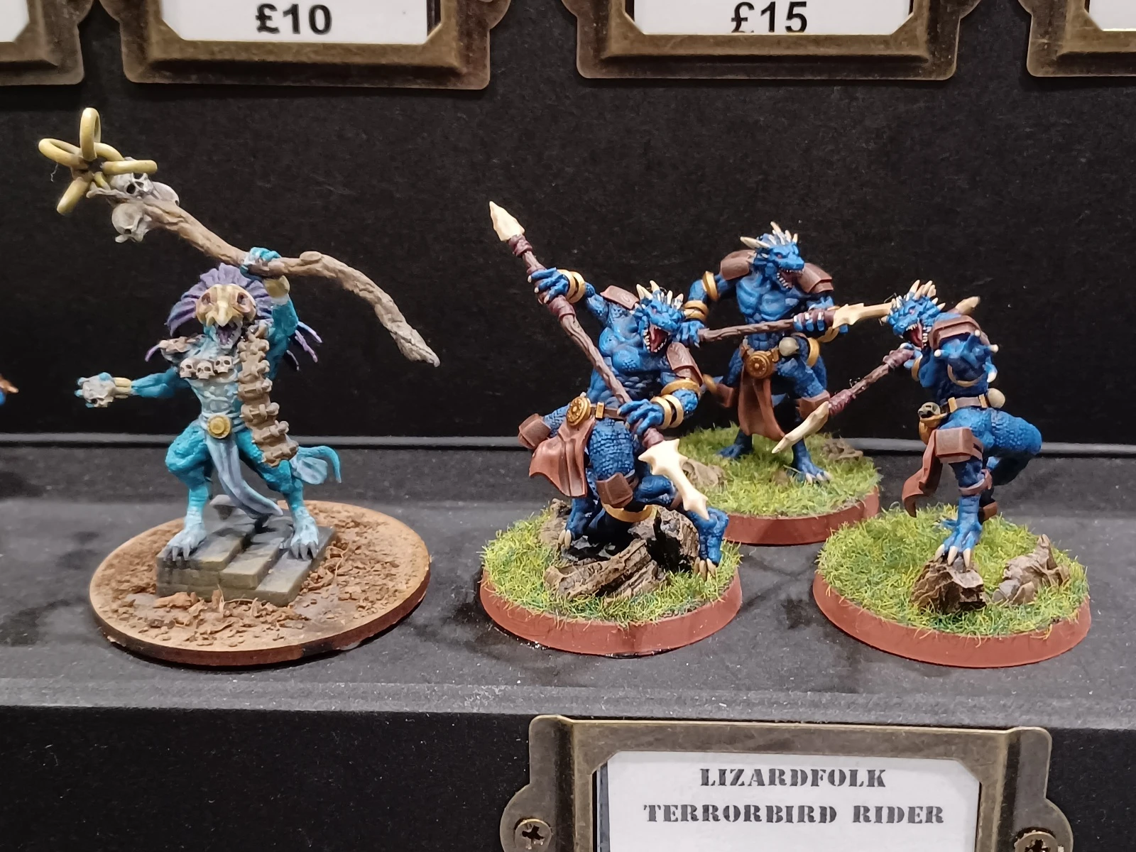 A group of four lizardfolk miniatures, each painted in vibrant blue with detailed scales and features, are displayed on a dark background. They are equipped with spears and wear brown leather armor, standing on circular bases adorned with grass and small rocks. A sign below them reads