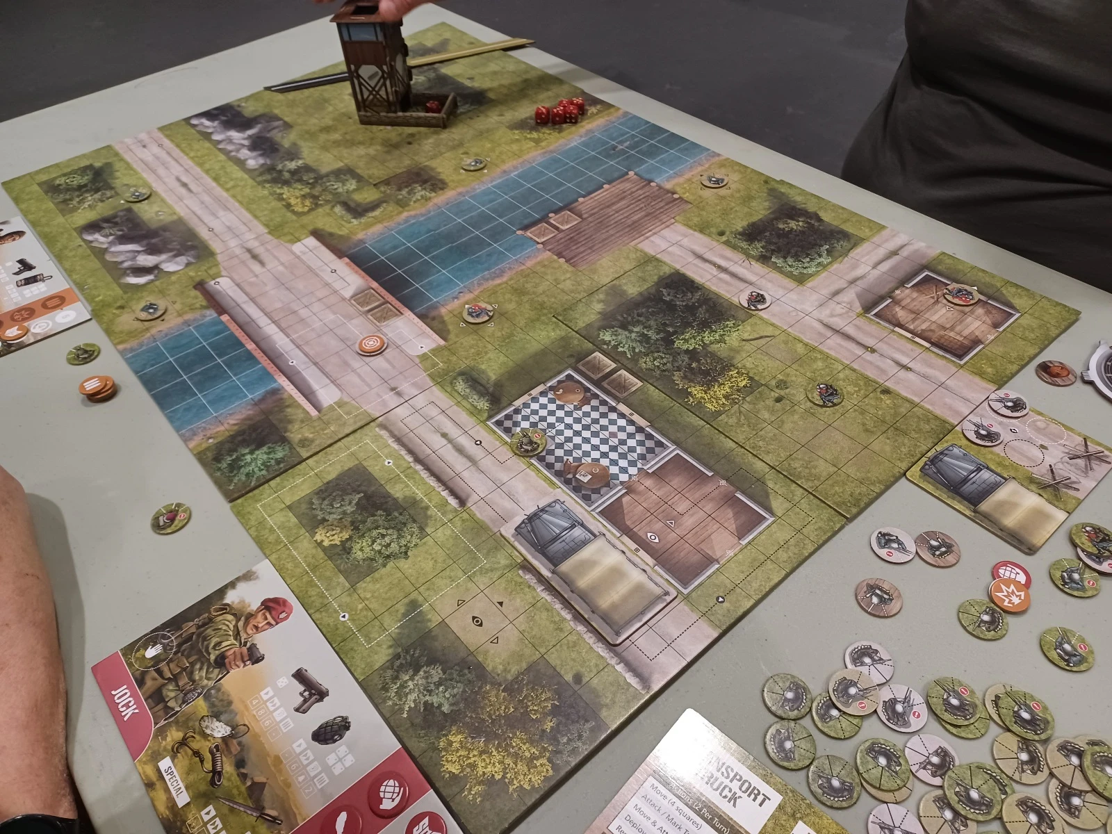 A tabletop gaming scene featuring a detailed game board set in a grassy area with a small water feature. The board includes various terrain elements such as houses, trees, and pathways, with miniature figures positioned throughout. Nearby, game tokens and a rule sheet are visible, indicating gameplay mechanics and actions.
