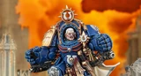 A detailed miniature of Marneus Calgar, a prominent character from the Warhammer 40,000 universe, is depicted in a striking pose. He wears ornate blue power armor adorned with gold embellishments, featuring a prominent Ultramarines symbol on his chest and a skull motif. The background is a dramatic orange, suggesting a fiery battlefield, enhancing the intensity of the scene.