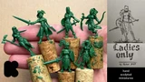 A hand holding several green, unpainted miniatures stands atop cork bases, showcasing a variety of female fantasy characters, including warriors and rogues. Each figure is intricately detailed, with distinct poses and weapons, such as swords and bows. To the right, a sketch of a female warrior is displayed, accompanied by the text "Ladies Only" and "hand sculpted miniatures" at the bottom.