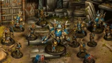A group of Cygnar miniatures is depicted in a detailed tabletop setting, showcasing the Gravedigger Auxiliaries unit. In the foreground, a character heavy warjack named Beaker stands prominently, adorned in blue and gold armor, with various mechanical features. Surrounding it are several elite veterans, including a female commander with red hair and a rifle, an ogrun captain, and a unit of Gravedigger Commandos, all featuring a mix of blue and gold uniforms, set against a backdrop of stone steps and a weathered door.