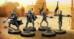 A group of four miniatures from the Star Wars universe is displayed against a desert backdrop featuring a simple structure. On the left, a character in a silver and black armor, resembling a bounty hunter, stands confidently. Next to him, two IG-Series Assassin Droids are posed with weapons drawn, showcasing their metallic bodies and intricate details. In the foreground, Grogu, a small figure with large ears, is depicted holding a small object, adding a whimsical touch to the scene. The ground is textured with grass and dirt, enhancing the overall aesthetic of the miniatures.