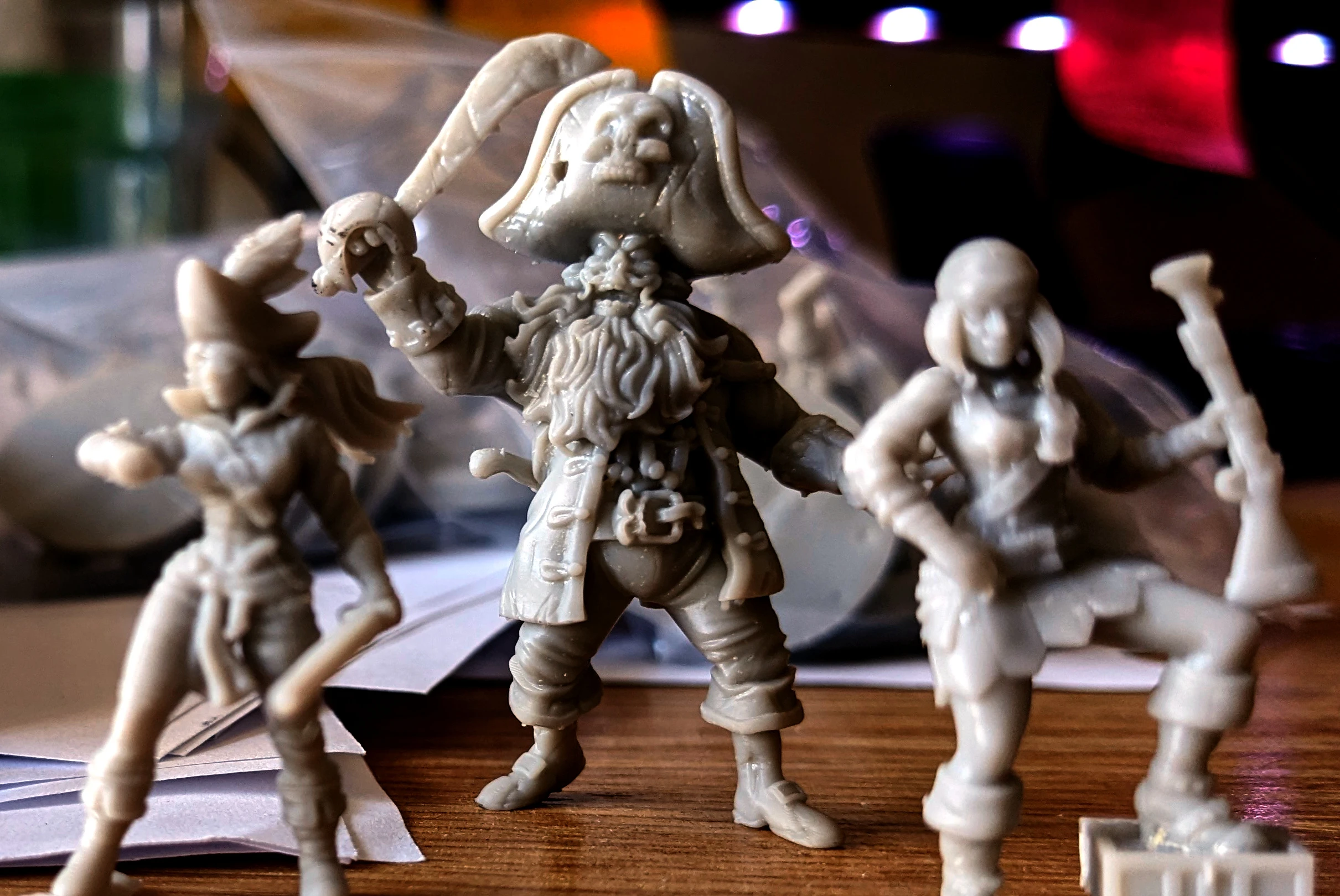 A collection of miniature pirate figures is displayed on a wooden surface, showcasing a variety of characters. In the foreground, a prominent pirate captain with a skull-adorned hat and a long beard holds a sword aloft, exuding a commanding presence. Beside him, a female pirate brandishes a cutlass, while other figures, including an undead pirate, are partially visible in the background, all rendered in a light-colored material that highlights their intricate details.